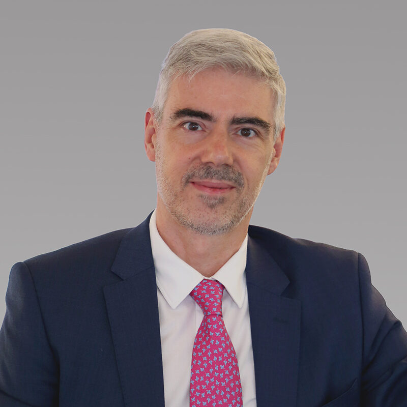 Javier Viejo leans up against a ledge. His smile is friendly and open. A bright pink patterned tie complements his expression.